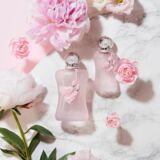 Parfums de Marly Delina La Rosee getoond in twee maten. Roze parfumsflesjes met kwastjes en pioenrozen eromheen liggend