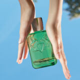 Parfums de Marly Greenley groen doorzichtige parfum fles wordt vastgehouden door twee hangen met een blauwe hemellucht